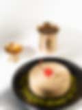 A round Indian sweet topped with a bright red cherry and cashews on a black plate sprinkled with crushed pistachios, with a brass lamp and cup in the background.