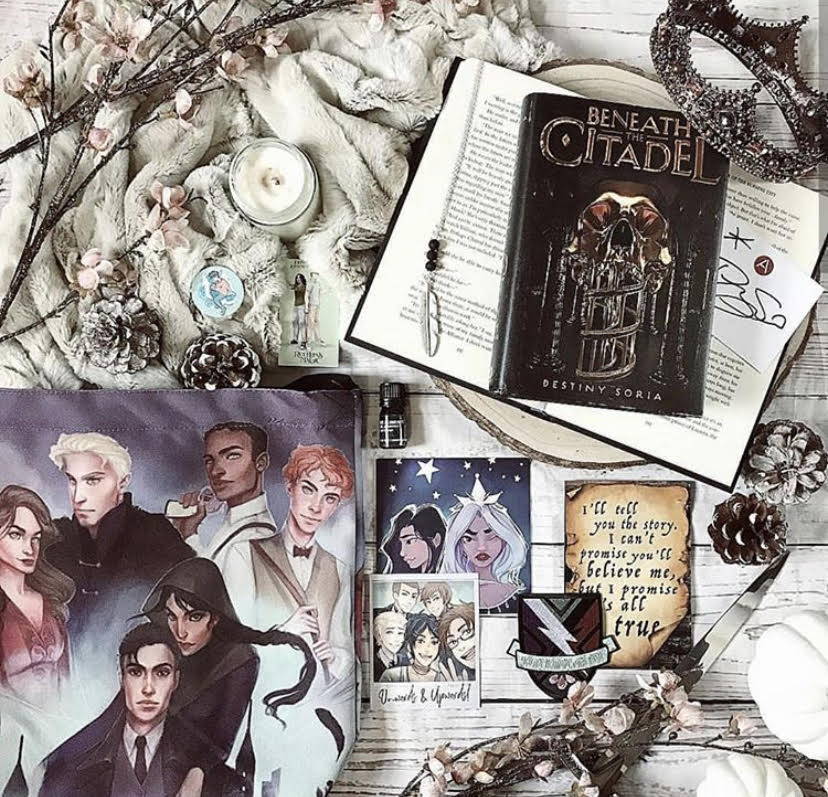 October 2018 Band of Misfits box theme included Beneath the Ciadel, E-Book Download of Ruthless Magic by @megancreweya, Adelina & Violetta Study Art Print, Those Aglionby Boys Essential Oil, Raven Cycle Essential Oil Diffuser Necklace, Roar Patch, Collectible Raven Cycle Polaroid, Six of Crows Crossbody Bag and Art Print, Percy Jackson Phone Stand and Sticker, and Double Sided Warcross Jersey Shirt included in Seelie and Solitary Fae boxes only. 