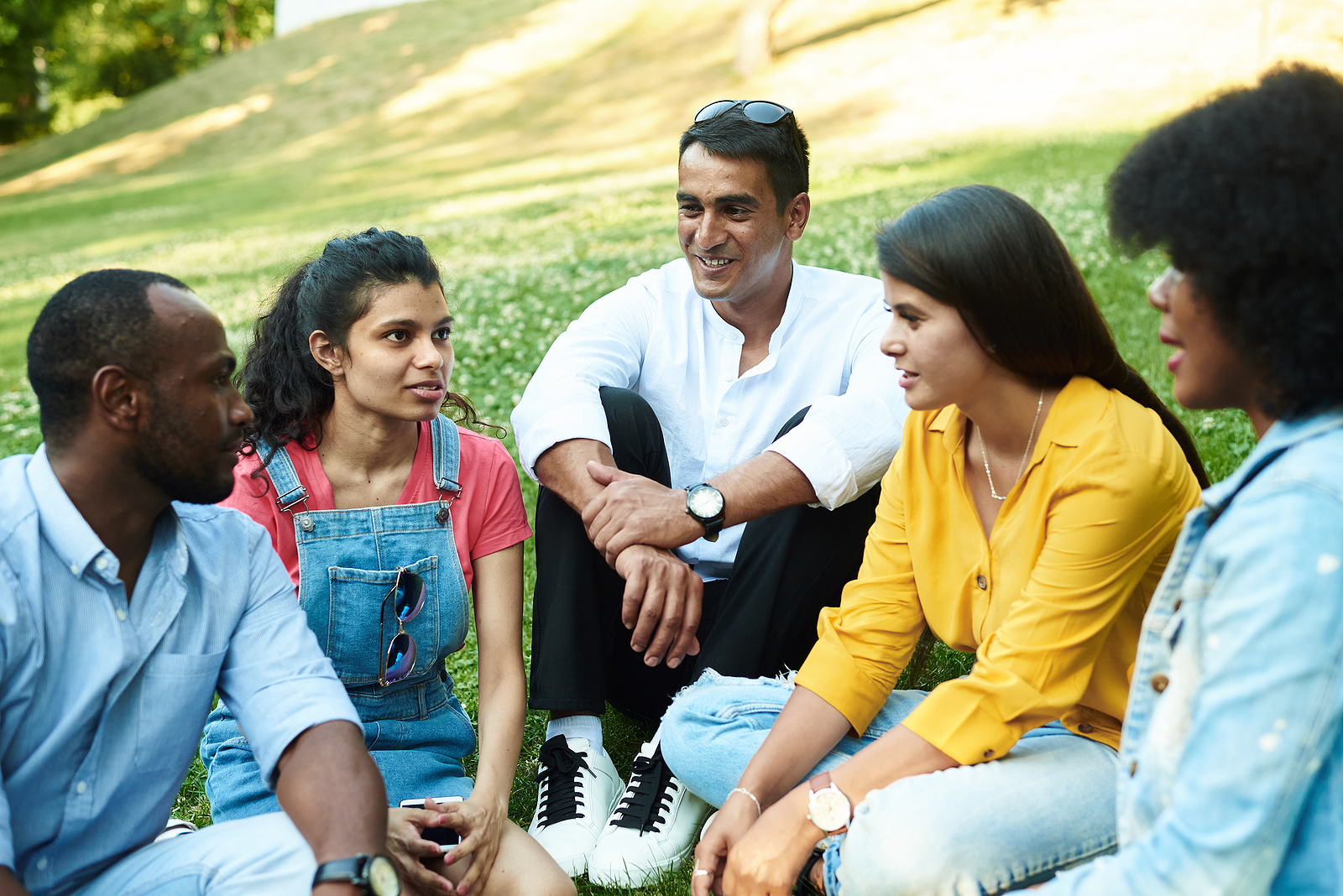 A group of mixed ethnicities and attractive friends chatting while sitting in the park on the grass wearing spring clothing.