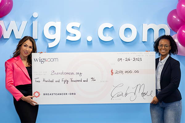 Two women are holding a large check in front of a wigs.com sign