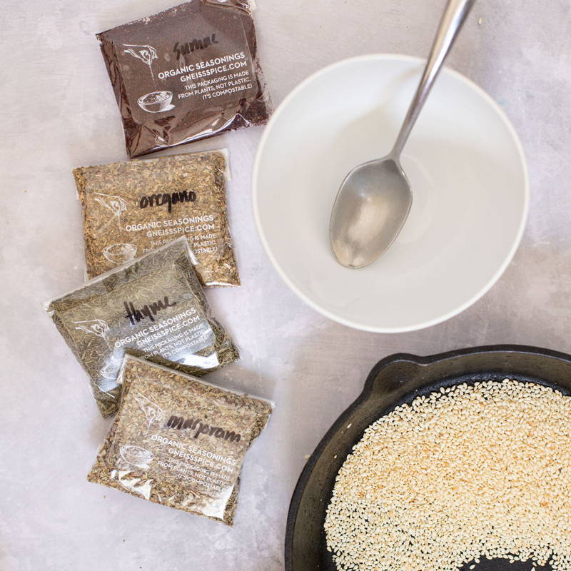 Four plastic spice pouches labeled 'Organic Seasoning' and the name of the spice it contains lay next to a white ceramic bowl and skillet full of sesame seeds