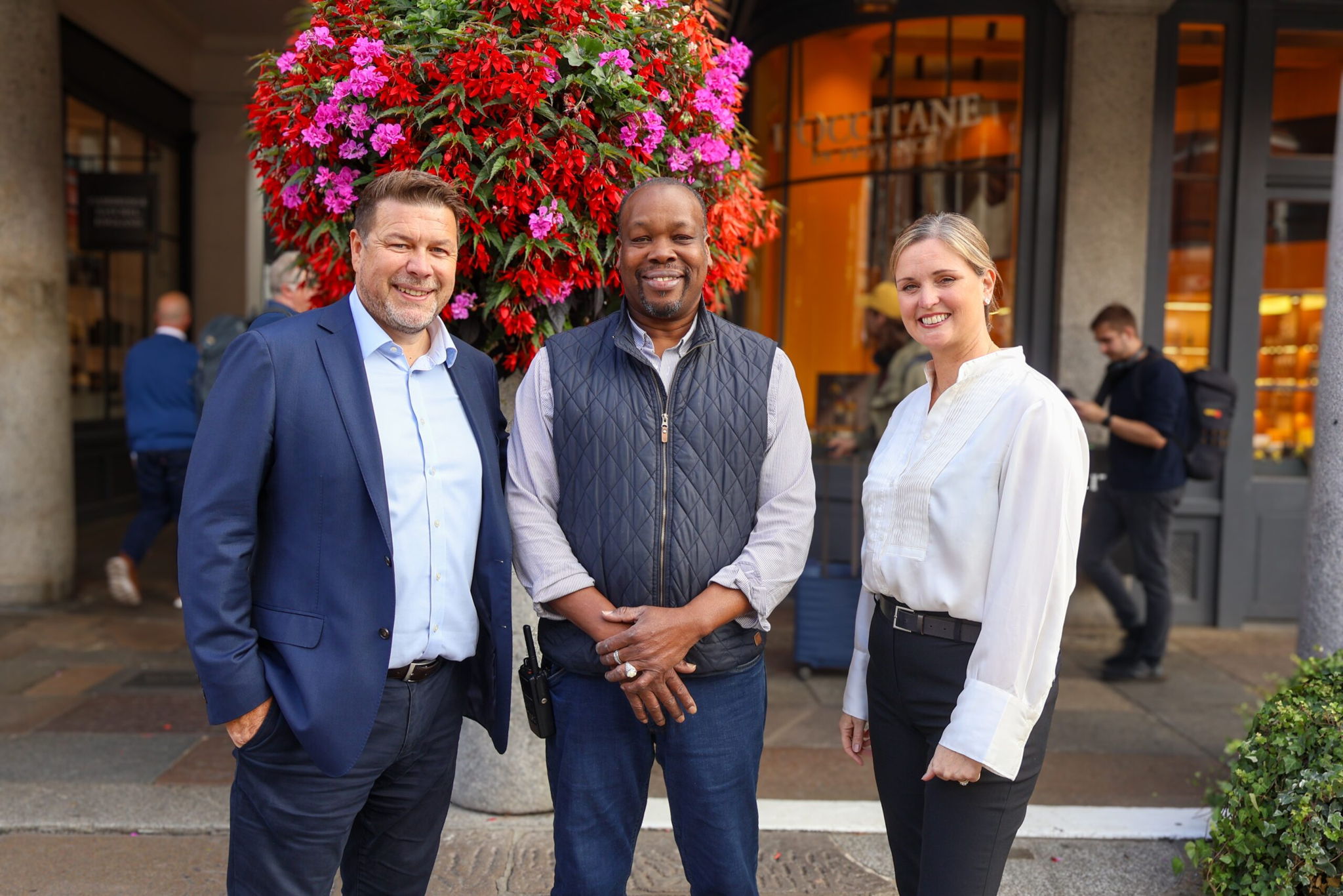 Three people stand smiling in front of a large planter of red and pink flowers on a city street, with shopfronts and pedestrians in the background. Two men wear business attire; one woman wears a white blouse.