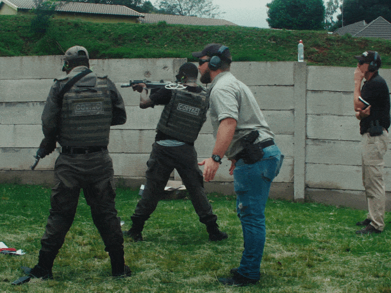 A man in a khaki shirt instructs two men in military gear who are shooting large guns.