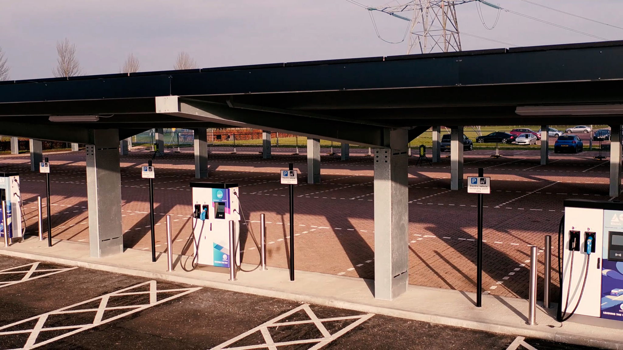 Electric vehicle charging station with multiple charging points under a large metal canopy, marked parking spots, and surrounding open area. Several cars are parked in the distance, with power lines visible in the background.