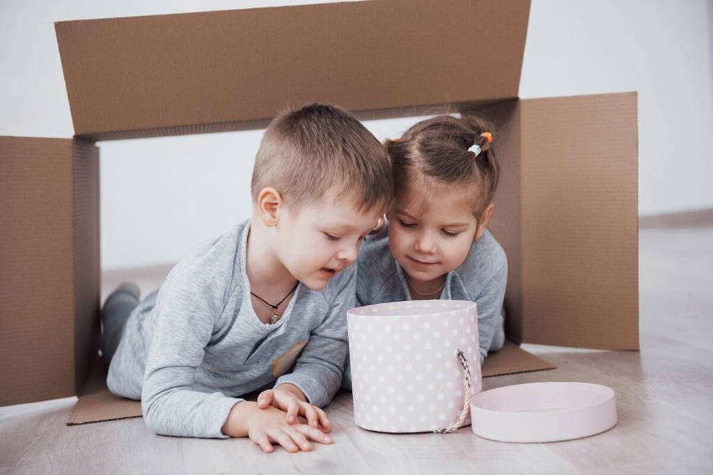 Deux jeunes enfants en pyjamas gris allongés sur le sol à l'intérieur d'un fort en carton, regardant curieusement dans une boîte-cadeau à pois roses.