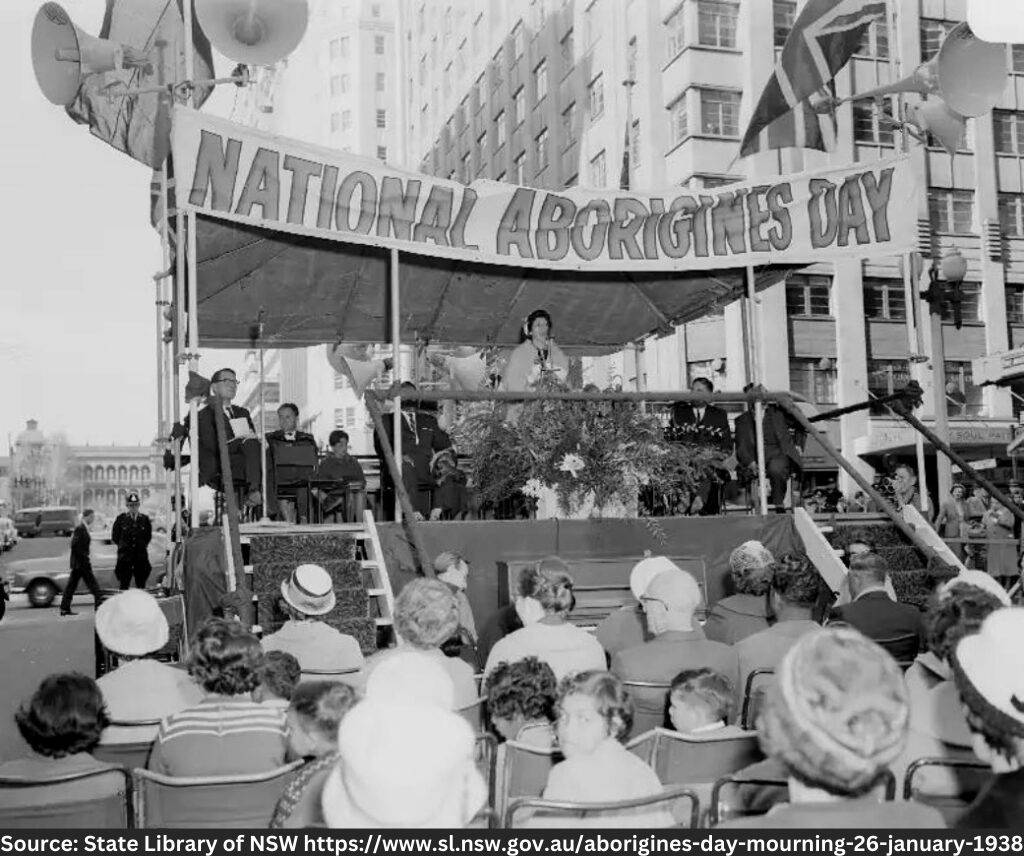 Black and white image of woman stood stage in front of an audience with banner titled 'National Aborigines Day'