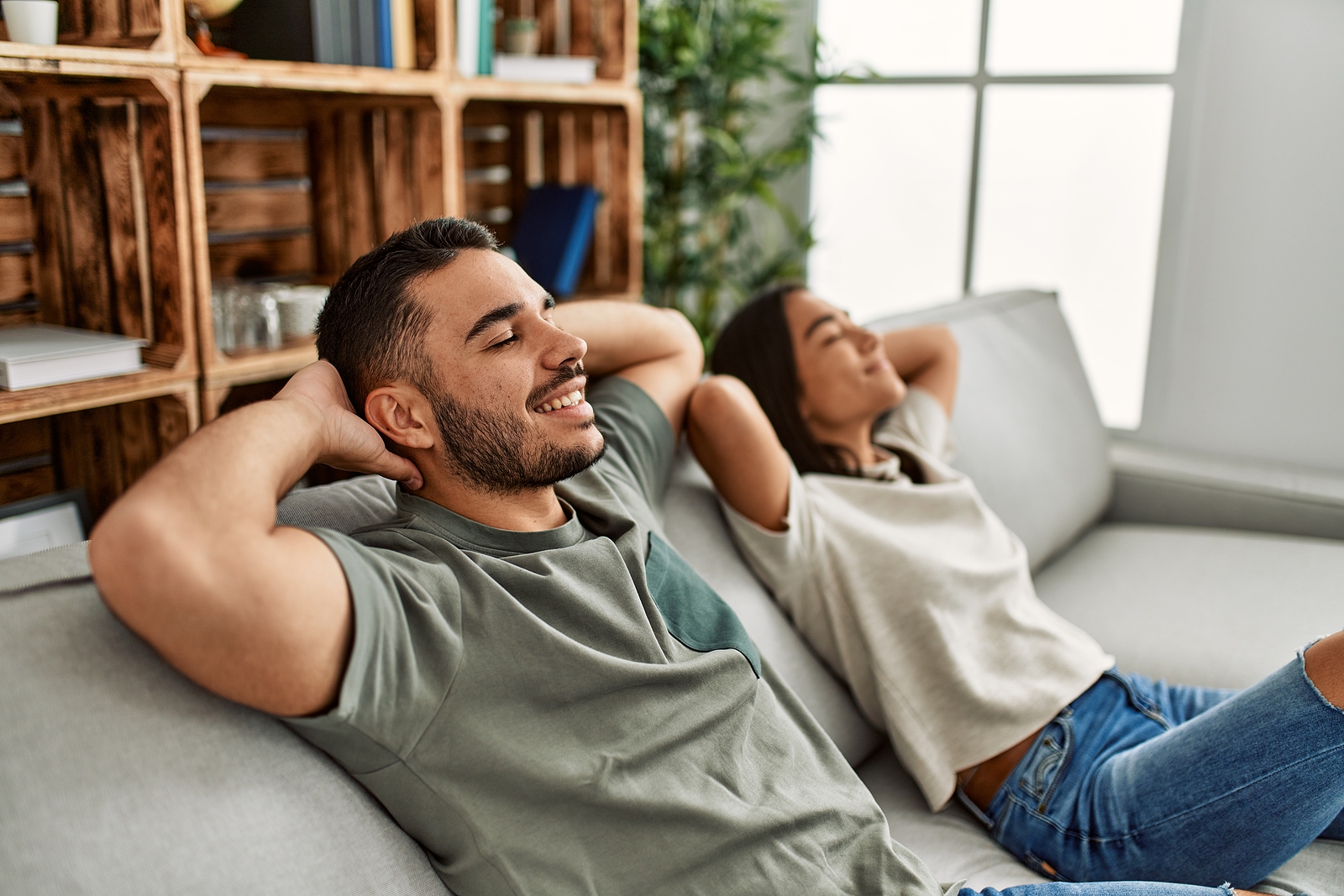 An attractive latino man and woman lay on the couch while reclining and their arms behind their heads with eyes closed.