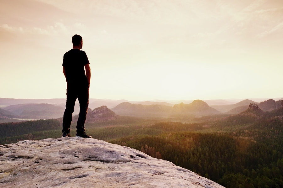 A man is standing on the edge of a cliff overlooking the rising sun over the valley.