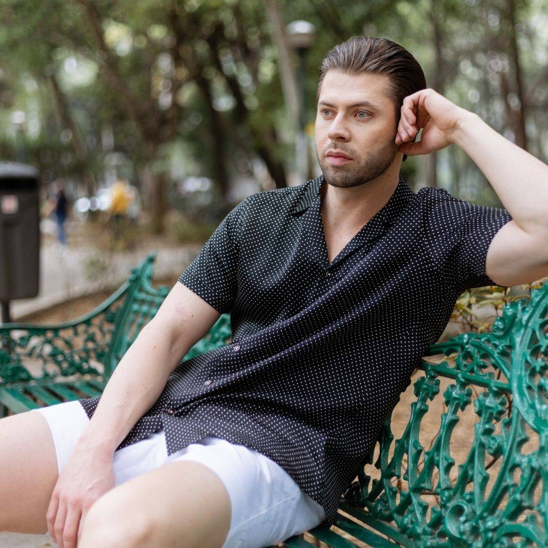 model sitting on a bench in a park wearing white shorts and a short sleeve black dotted silk shirt from 1000 kingdoms