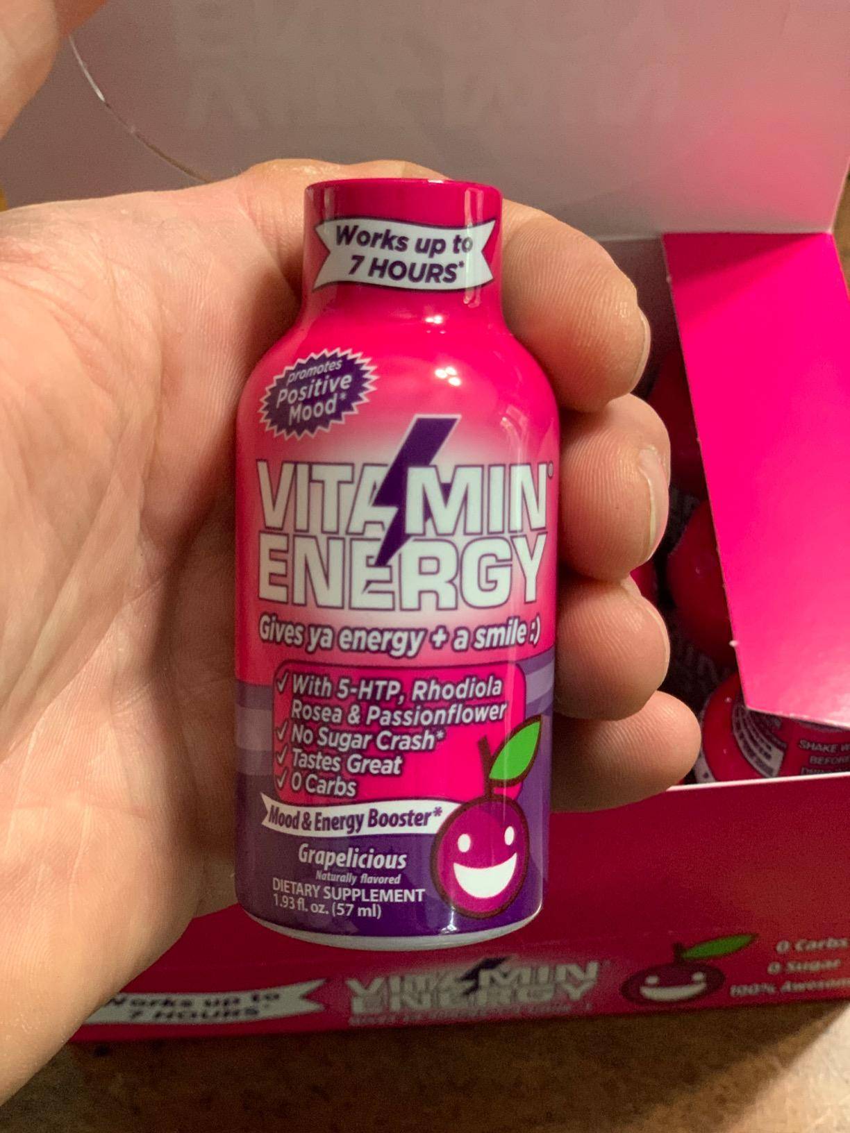 Person holding a bottle of Vitamin Energy drink, labeled 'Grapelicious,'promising energy and a positive mood.