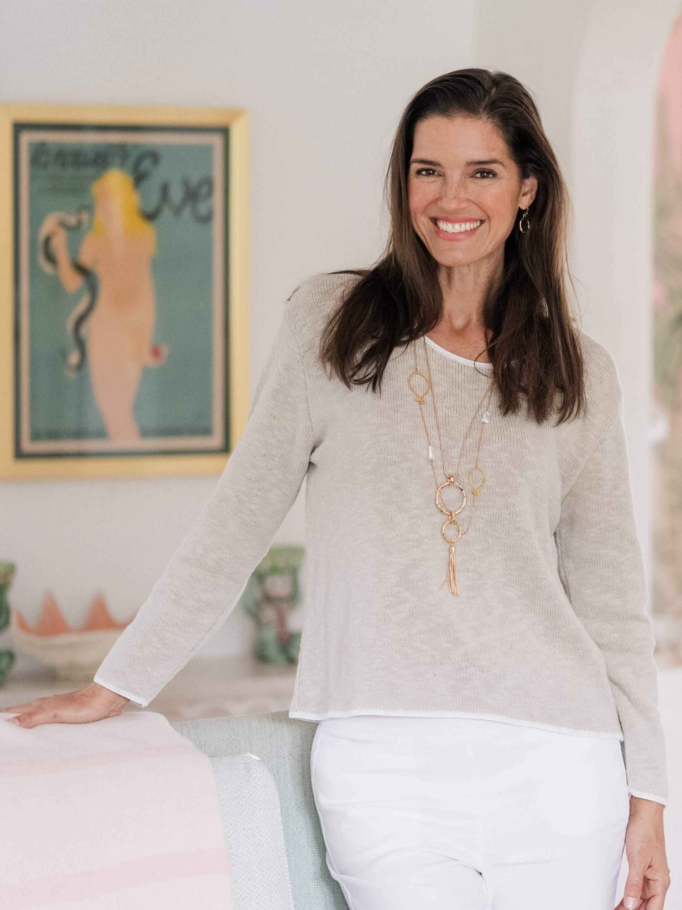 A woman in a beige sweater and white pants leans against a sofa with a painting on the wall in the background