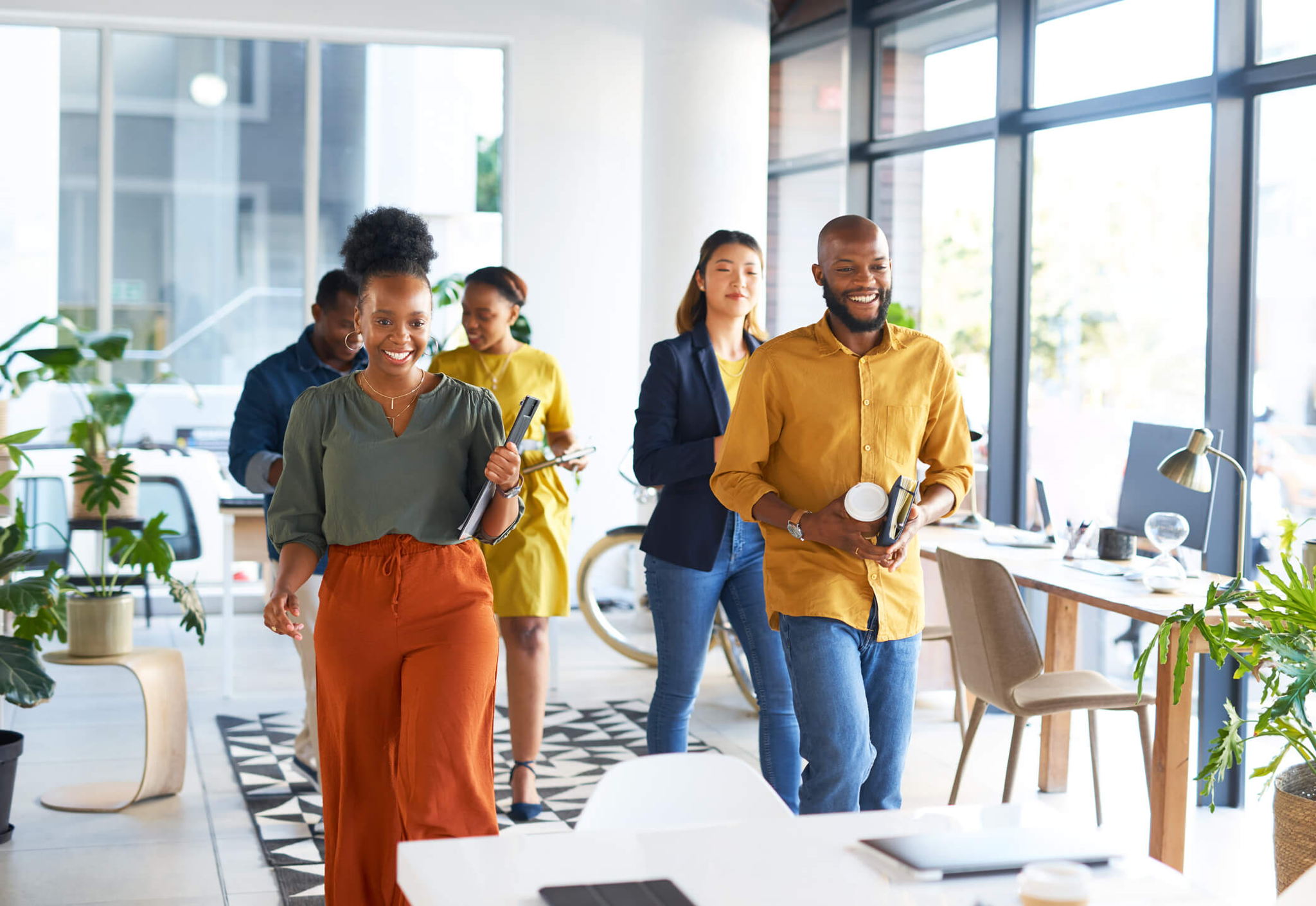 A diverse group of five people walks through a modern, well-lit office space. They seem cheerful and are holding various items like books and coffee cups. The office features large windows, plants, and an inviting, open layout.