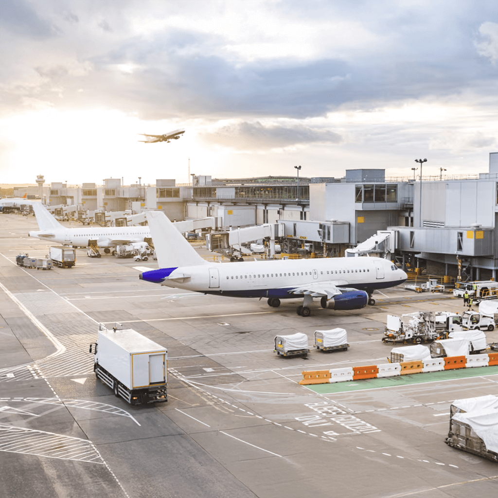 Commercial airplanes parked at airport gates with jet bridges attached, ground service vehicles nearby, and another plane taking off in the background under a partly cloudy sky at sunset.