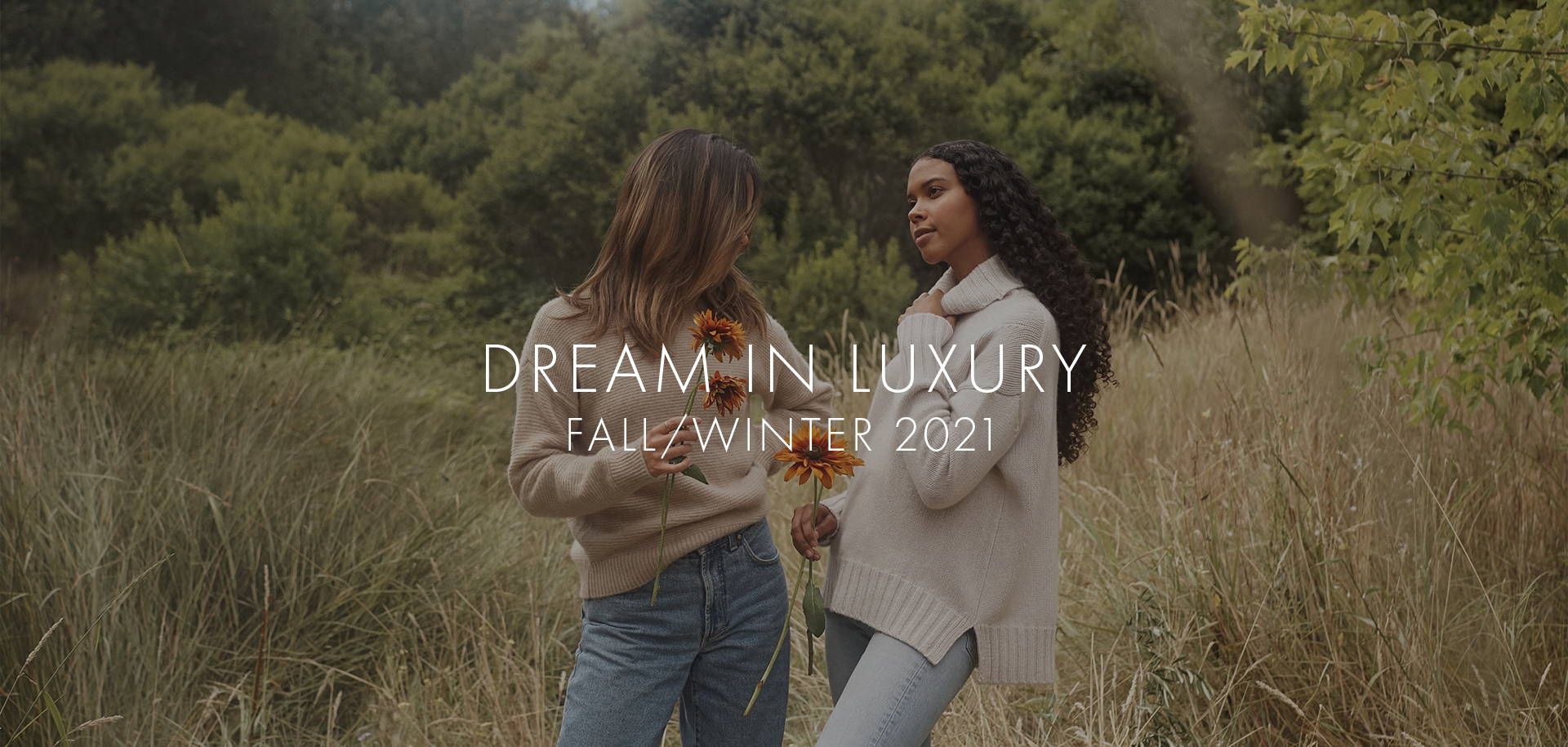 Dream in Luxury, Fall/Winter 2021 text on an image of two models wearing a beige pullover and a grey pullover, standing in bushes and holding flowers. 