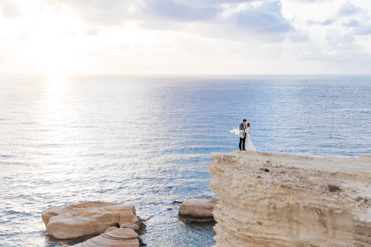 Un couple de mariés se tenant sur les falaises de Paphos, en robe de mariée et costume, la mer bleue en arrière-plan, photographie de mariage cinématographique, prise de loin.