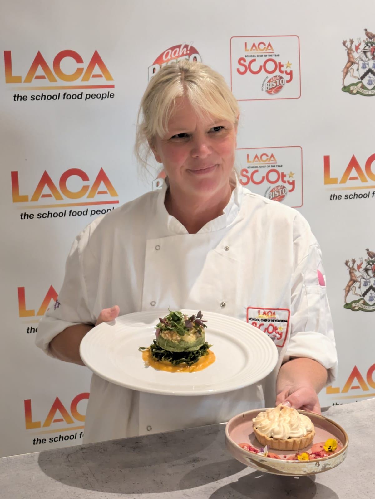 A smiling chef in a white uniform holds two plated dishes, one with a savory layered dish and another with a tart, standing in front of a backdrop with LACA and Scoff Bistro logos.