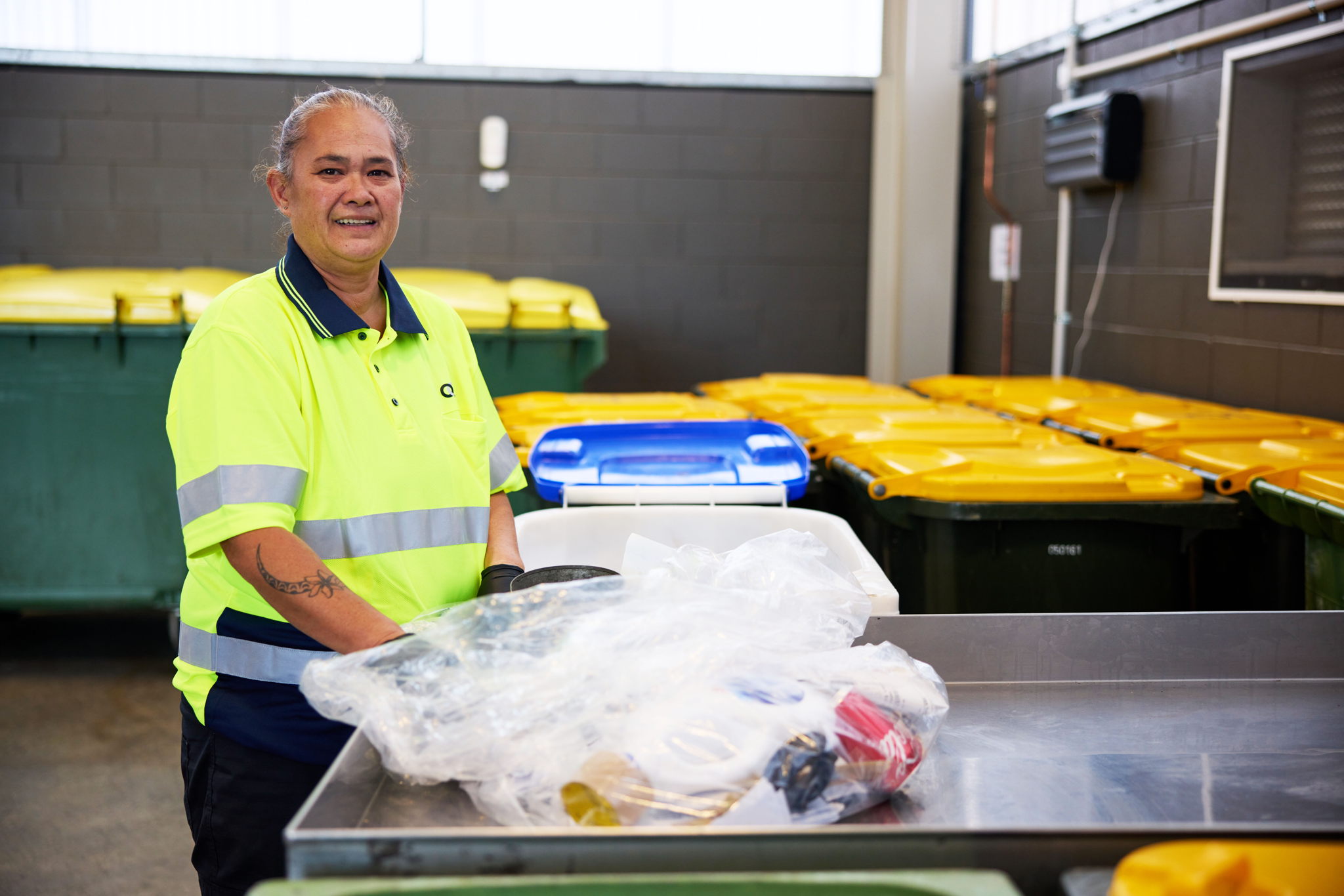 A person wearing a high-visibility yellow shirt stands indoors beside a sorting table with plastic waste, surrounded by large recycling bins. The person is smiling and appears to be engaged in recycling work.
