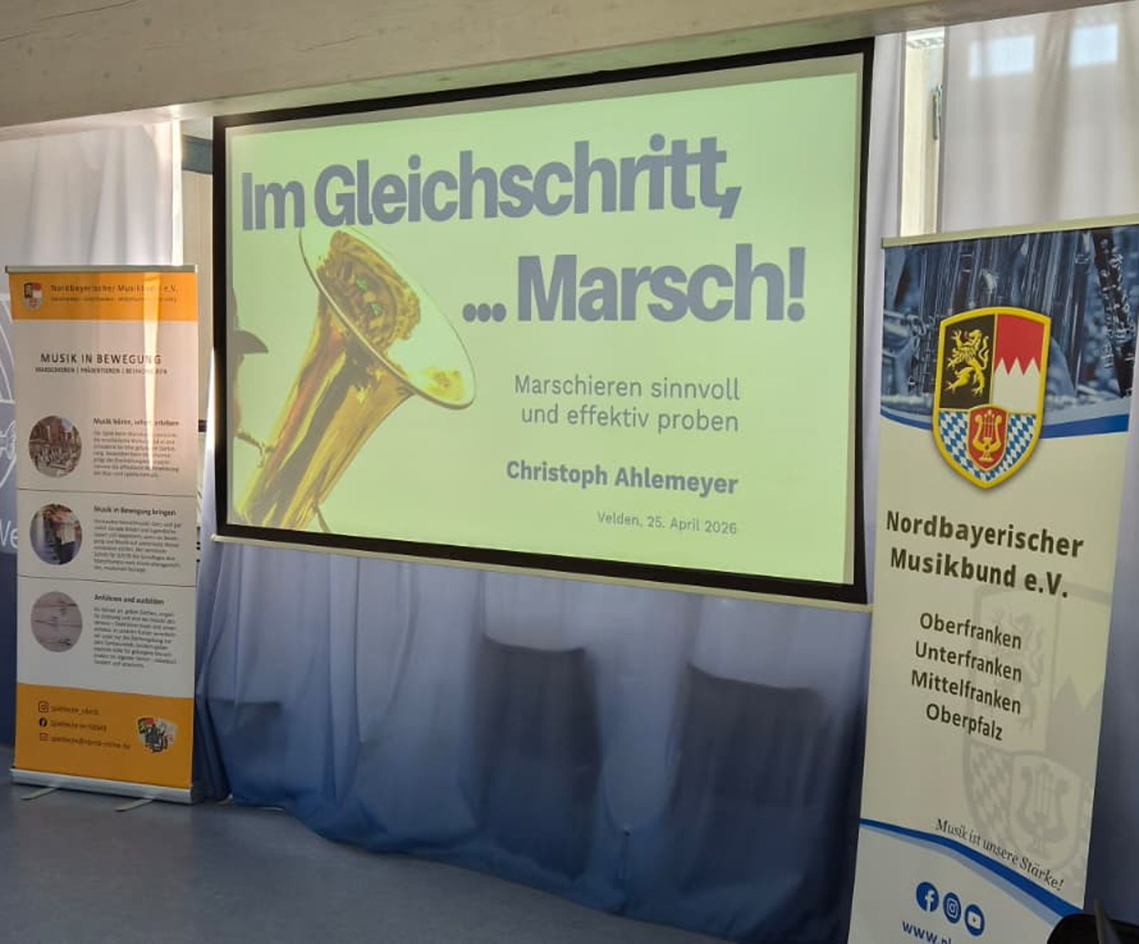 Neben einer Leinwand stehen zwei Roll-ups. Auf der Leinwand ist eine Präsentation zu sehen mit der Aufschrift "Im Gleichschritt, Marsch!".