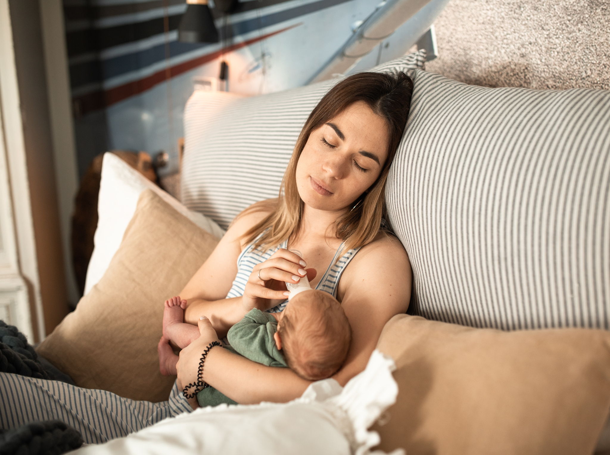 A young mom feeds her baby with a bottle while she closes her eyes.