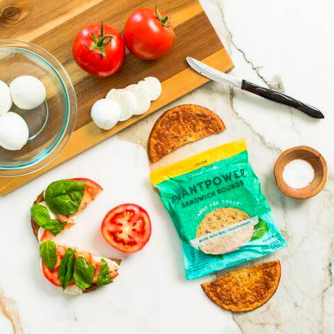 Tomatoes, basil, mozzarella cheese, and Outer Aisle Sandwich Rounds on a cutting board and a sandwich