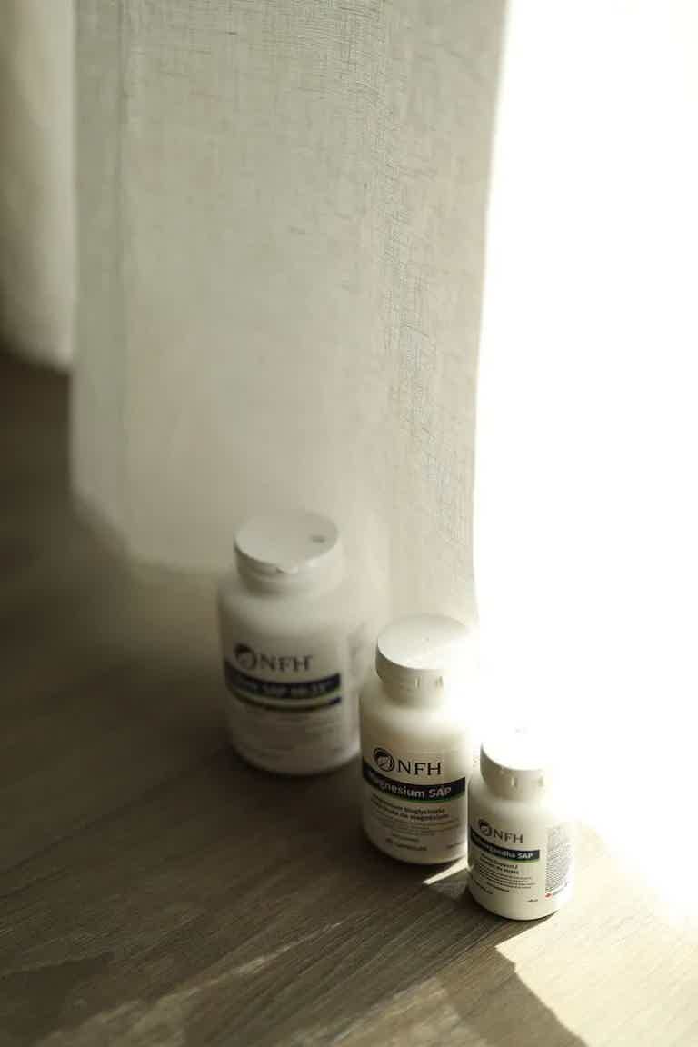 Three white supplement bottles on a wooden floor next to a sheer curtain with soft natural light.