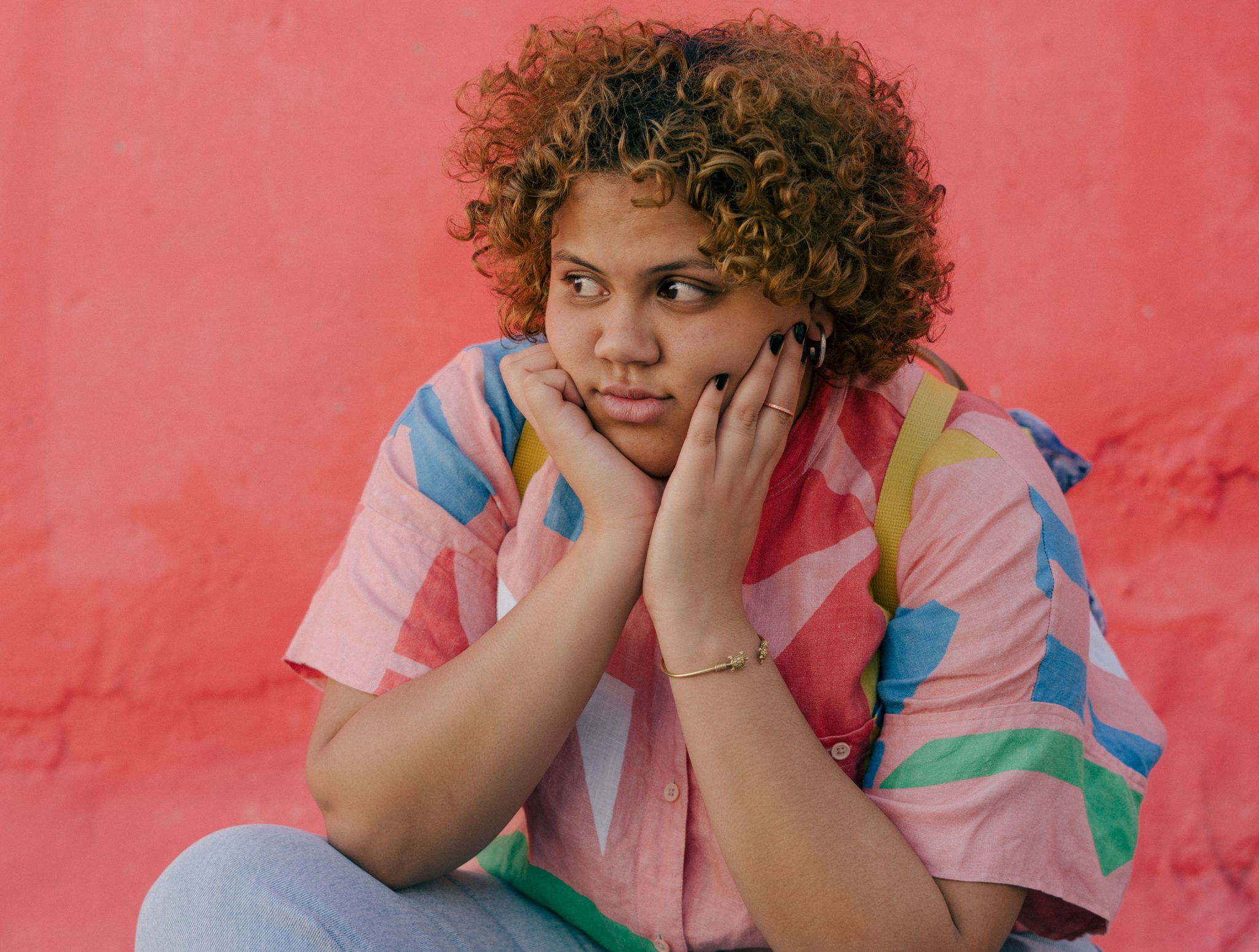 A young black woman woman with curly hairs sits with her hands on her chin looking to the side.