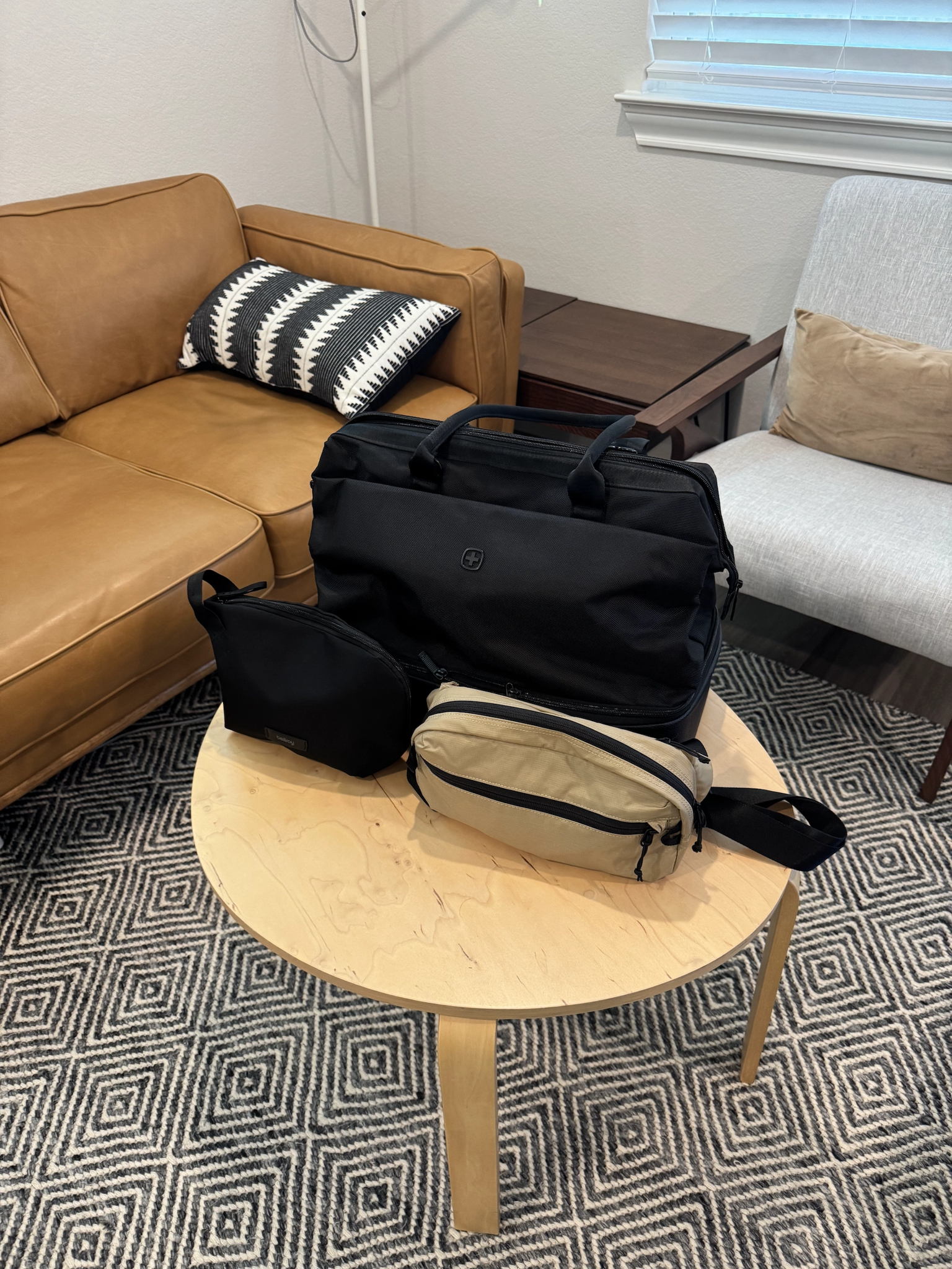 Weekender bag with the top compartment open, showing a Tomtoc sling bag and a small camera pouch neatly packed inside. The bag rests on a tidy bed in a softly lit room, creating an organized and calm atmosphere. No visible text in the image.