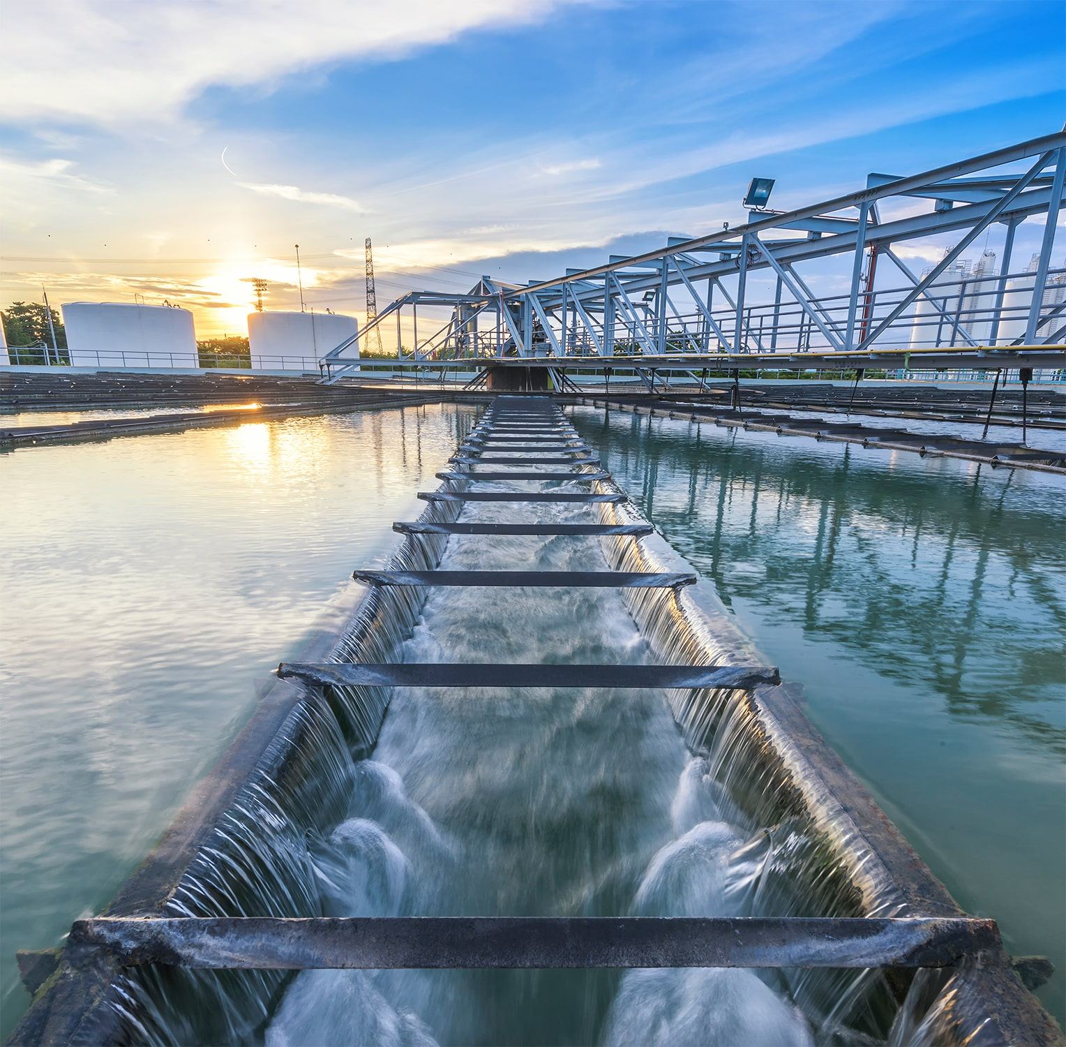 A modern water treatment facility at sunset, showing clear water flowing through a series of channels with metal structures and large storage tanks in the background.