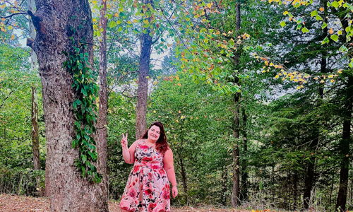 Person in a floral dress poses with a peace sign beside a tree in a vibrant, serene forest, exuding joy and tranquility.