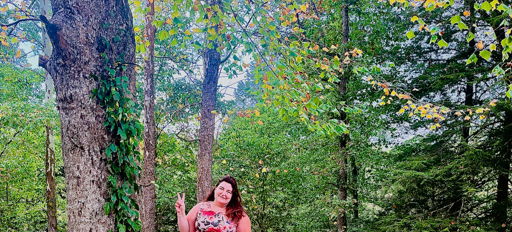 Person in a floral dress poses with a peace sign beside a tree in a vibrant, serene forest, exuding joy and tranquility.