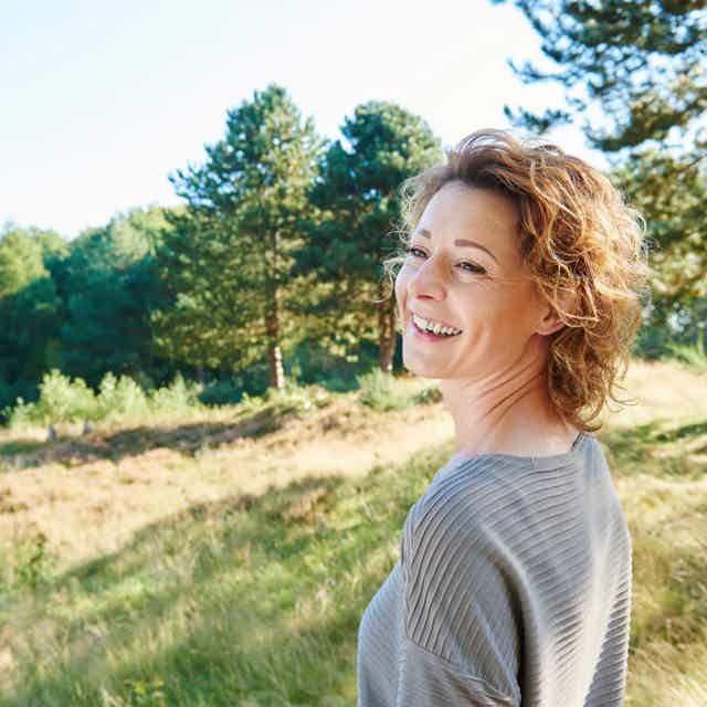 Smiling woman with short curly hair stands in sunny meadow with trees, wearing a gray ribbed top.
