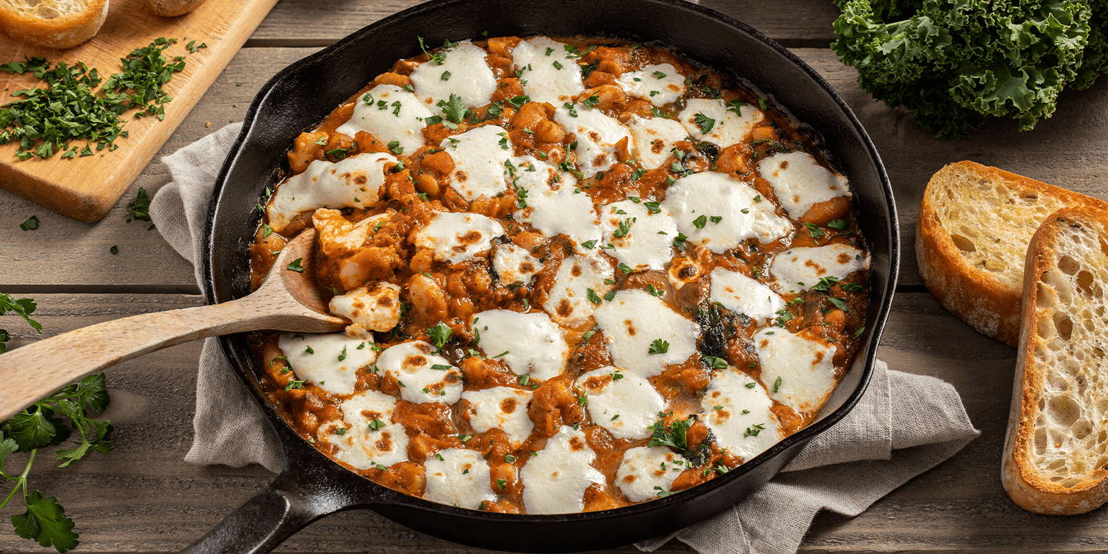 A wooden tabletop with a cast iron skillet of the prepared ZENB Tomato And White Bean Skillet recipe sitting on a gray cloth and styled with a wooden serving spoon, whole ingredients, and sliced bread on the side.