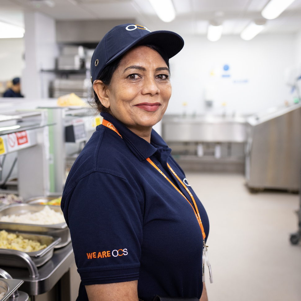 A smiling woman in a navy uniform and cap stands in a commercial kitchen. She wears a lanyard and the shirt logo reads WE ARE OCS. Food trays and kitchen equipment are visible in the background.