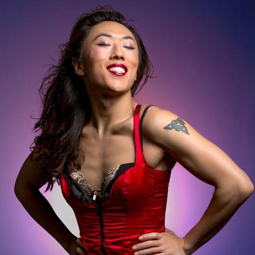Nicky wearing a red corset and her hands on her waist smiles with eyes closed against purple background.