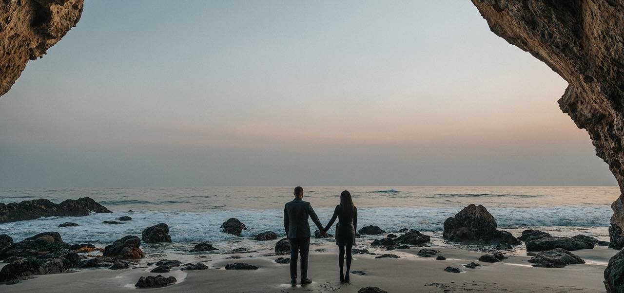 Paar steht Händchenhaltend am Strand und schaut aufs Meer