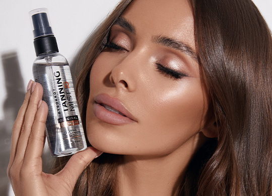 A beautifully bronzed girl holding the bottle of the Byrokko Tanning Mist.