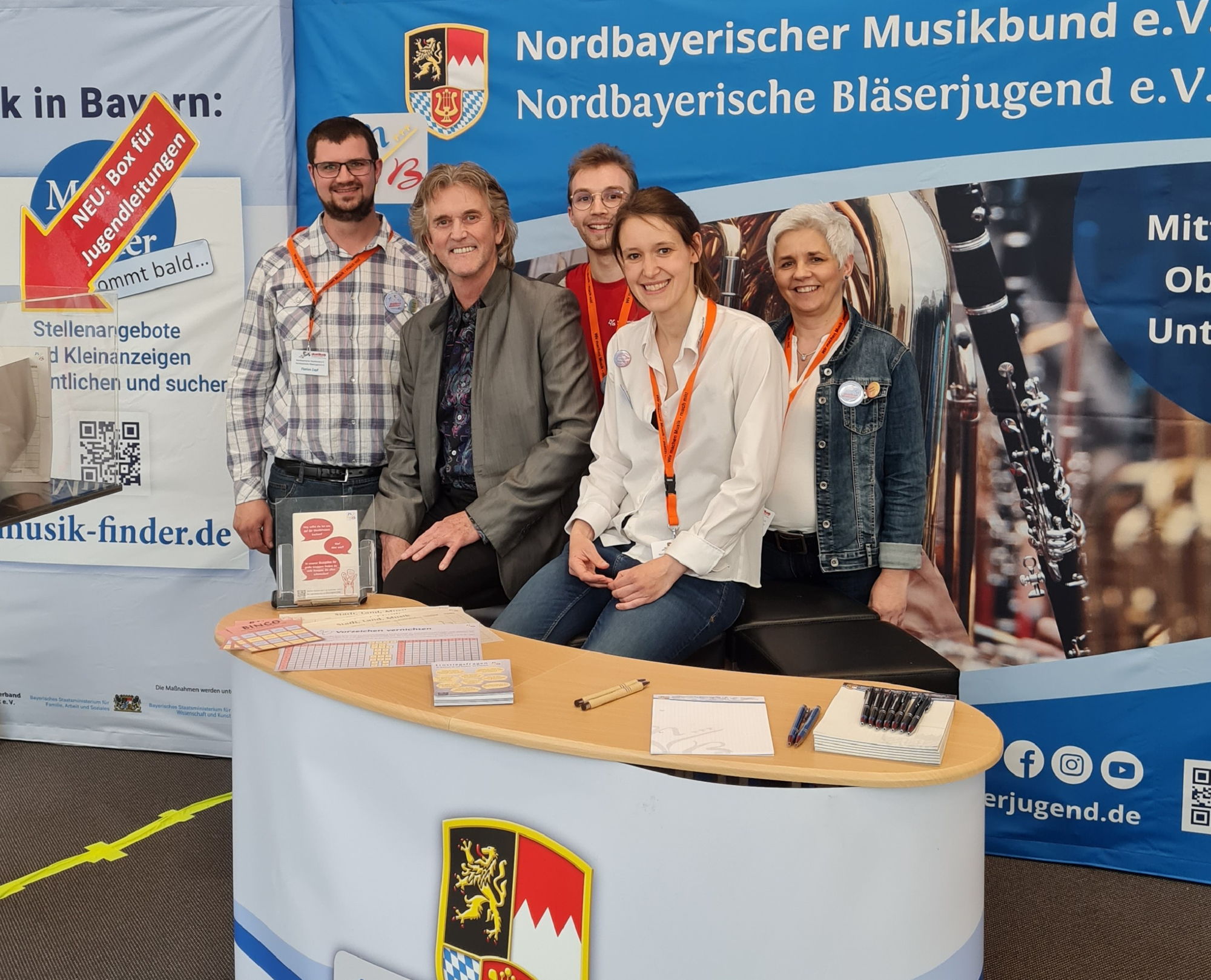 Drei Männer und zwei Frauen stehen hinter einer bunten Messetheke und lächeln in die Kamera. 