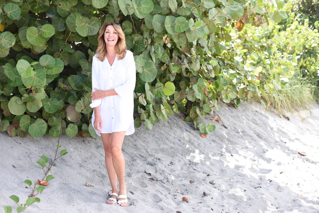 A woman wears a white button-front shirt cover-up on a beach.