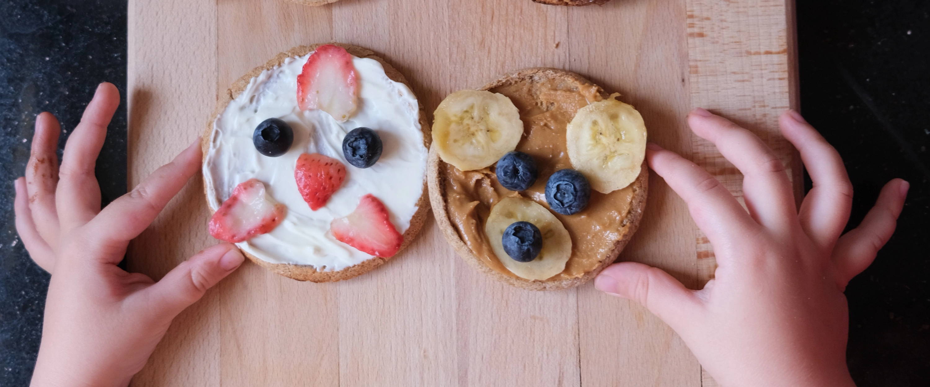 An overhead view of a wooden cutting board with four Pacha English muffins decorated as animal faces with spreads, berries, and bananas, with a child's hands reaching in to frame the bottom two snacks.
