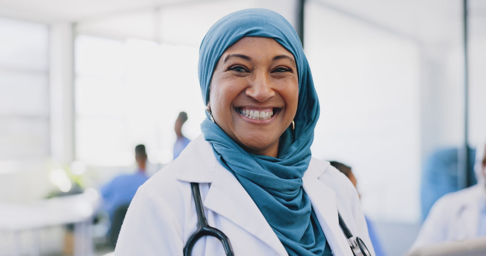A smiling woman wearing a blue hijab and a white lab coat with a stethoscope around her neck stands in a bright medical office. Other people in lab coats are blurred in the background.