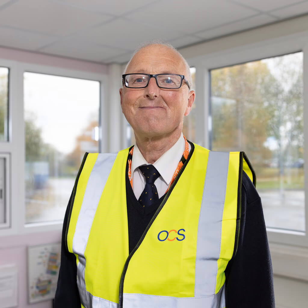 A specialist aviation services colleague wearing glasses and a high-visibility vest with the OCS logo stands indoors in front of large windows.