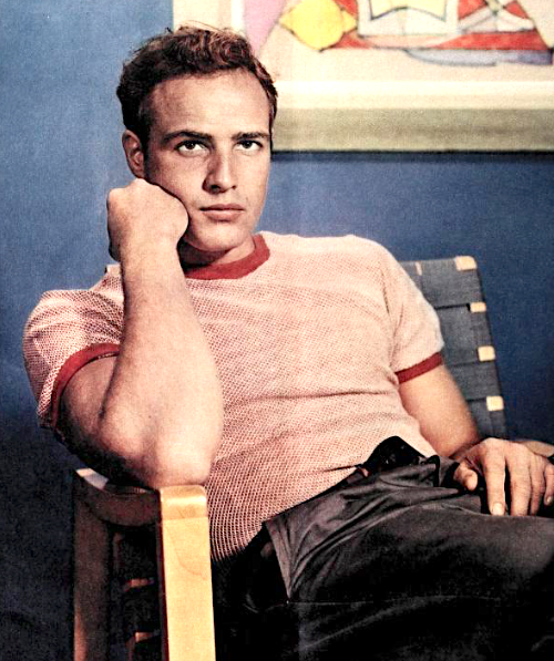 Marlon Brando sitting on a chair wearing casual clothes, with an arm to his chin looking at someone out of frame.
