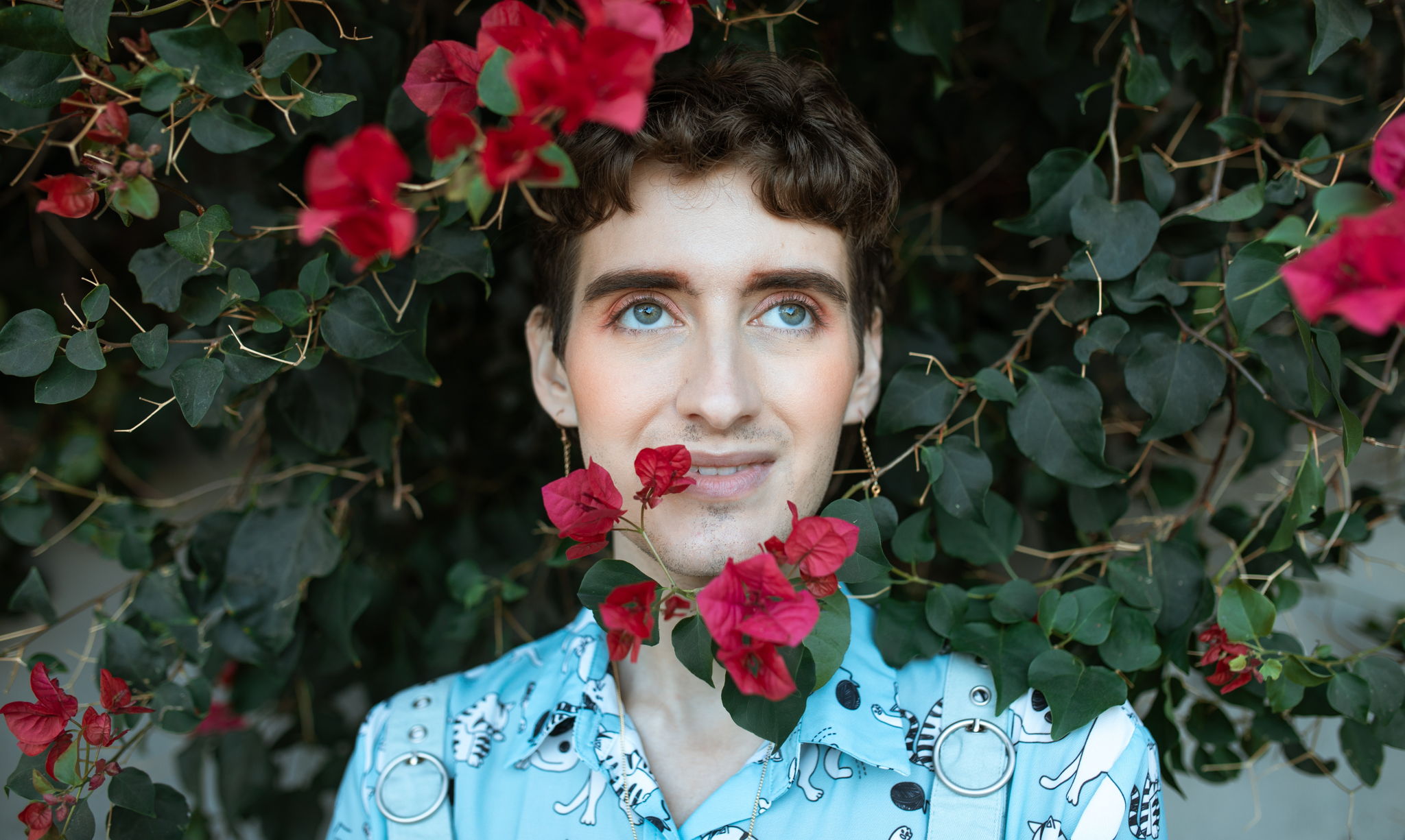 A non binary person smiles and looks to the sky in front of a large rose bush.