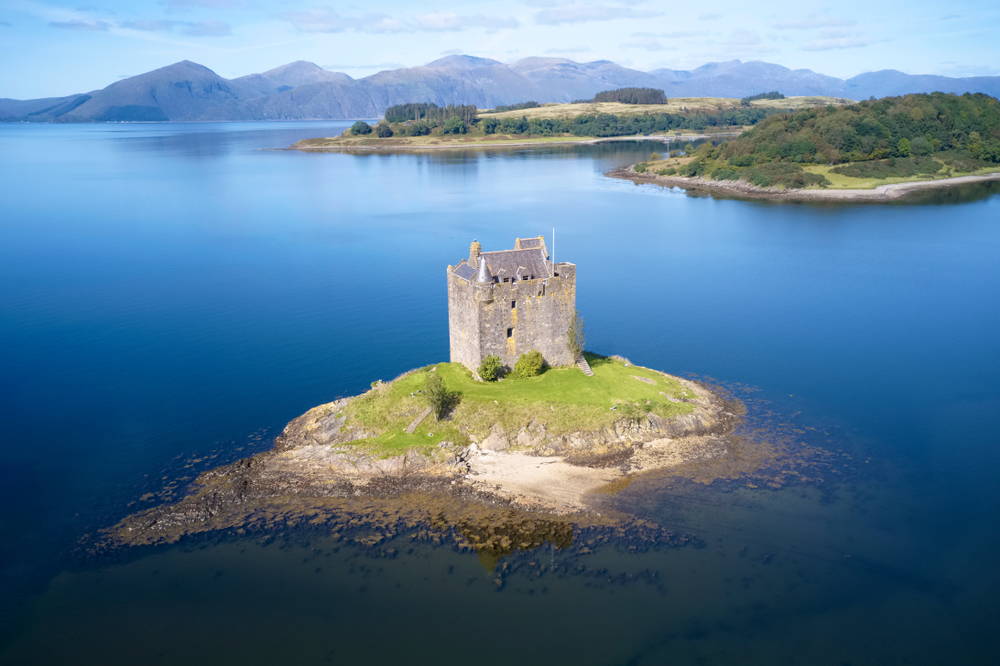 Castle Stalker siting on a small rocky island in the calm sea water of Loch Linnhe with the mountainous island of Mull in the distance.