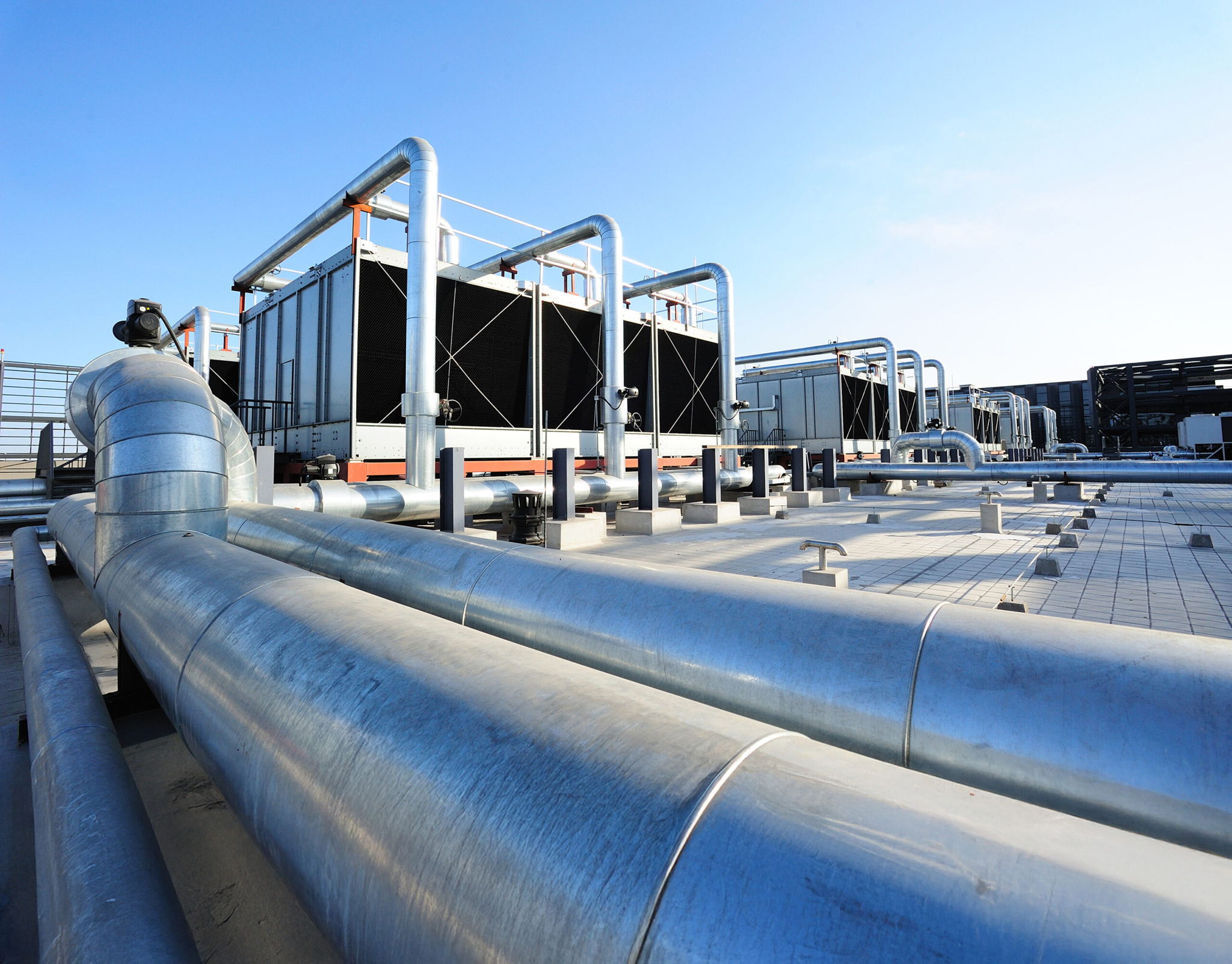 Large metallic pipes and industrial cooling units on a rooftop, set against a clear blue sky. The image suggests a modern HVAC or industrial refrigeration system.