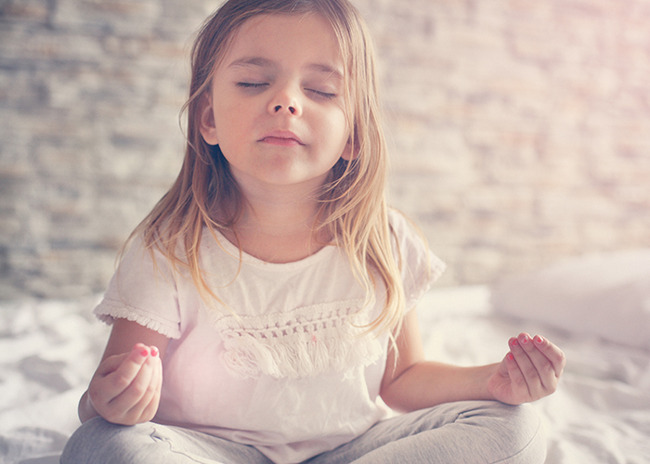 Little girl closes her eyes, sits cross legged and meditates on her bed