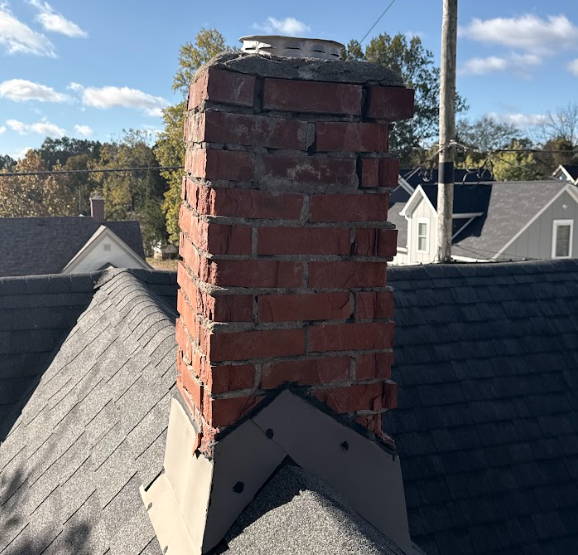 Brick chimney on a residential roof with deteriorating mortar joints and visible wear around the crown and flashing.