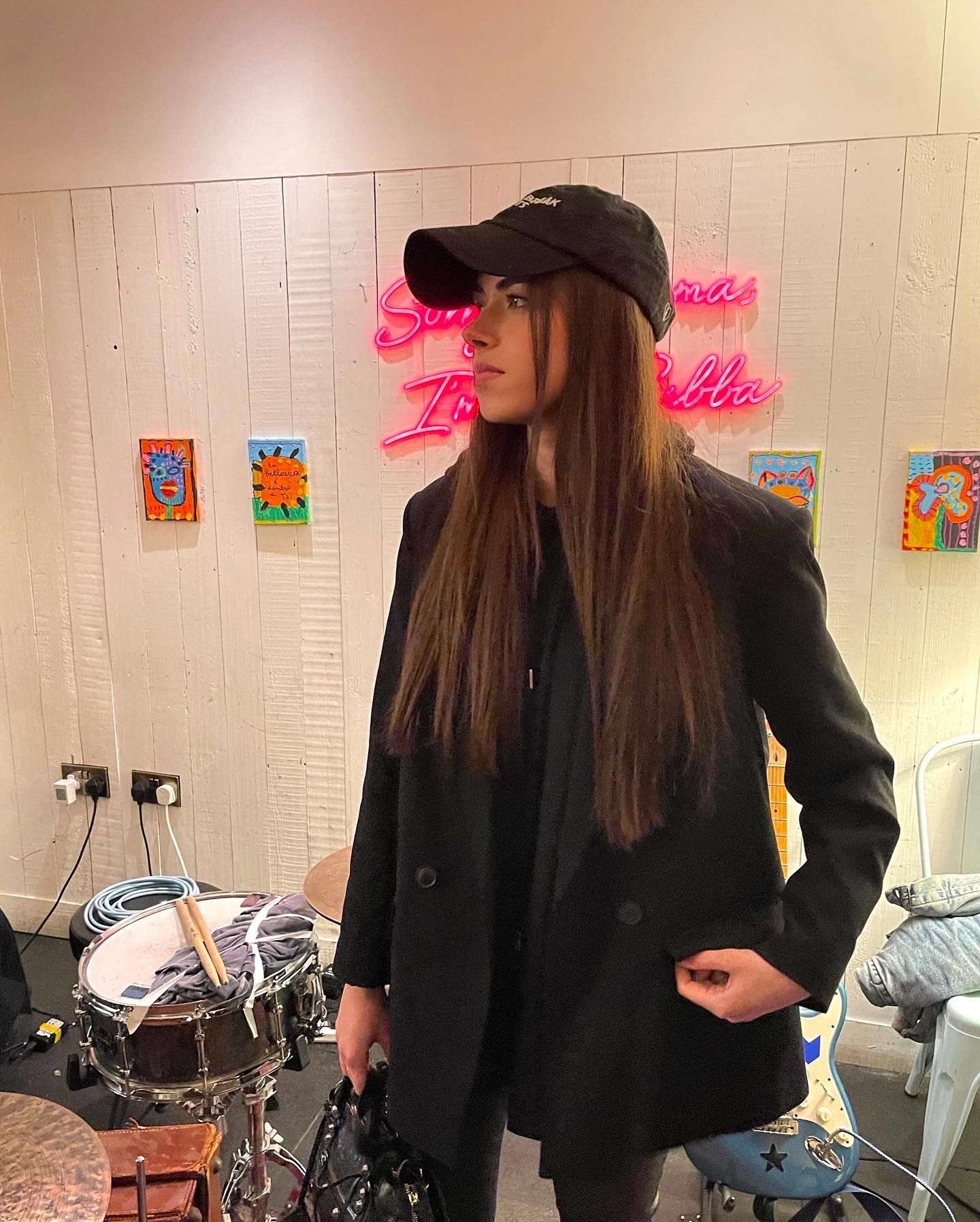 Stella standing in a black jacket with a black baseball cap in front of a wall covered in little painting. She is in front of a pink neon sign that we are unable to read. There is a drum with drumsticks in the photograph to her left.