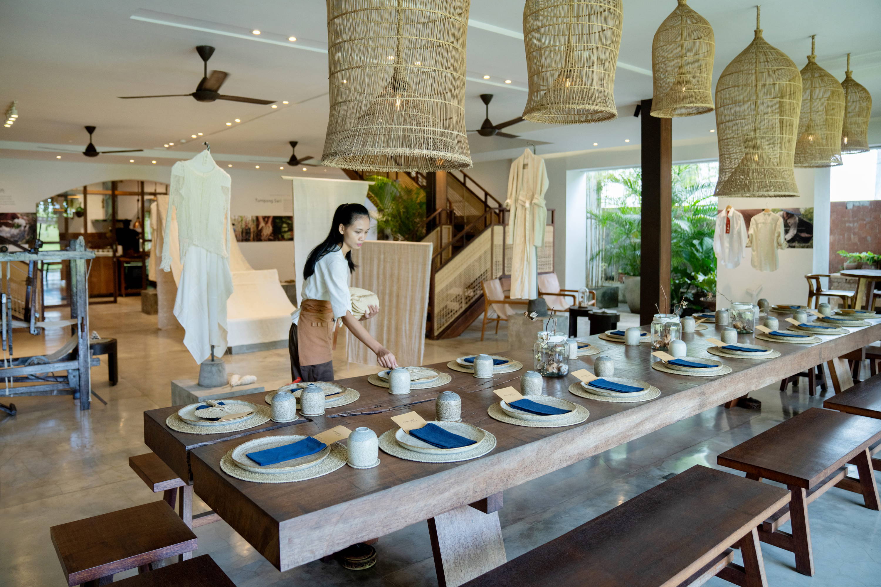 A woman is setting up tables with dinnerwares. Table is decorated with dried cotton plants. In the background, KAPAS Installation by SukkhaCitta in John Hardy Boutique & Gallery at Seminyak.