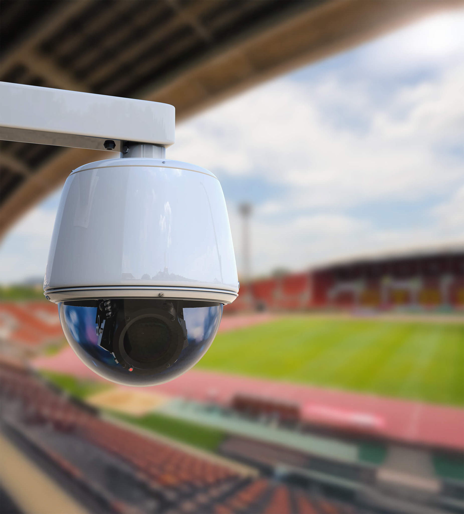 A close-up of a white security camera with a black lens, mounted outdoors, overlooking a blurred sports stadium with red seating.